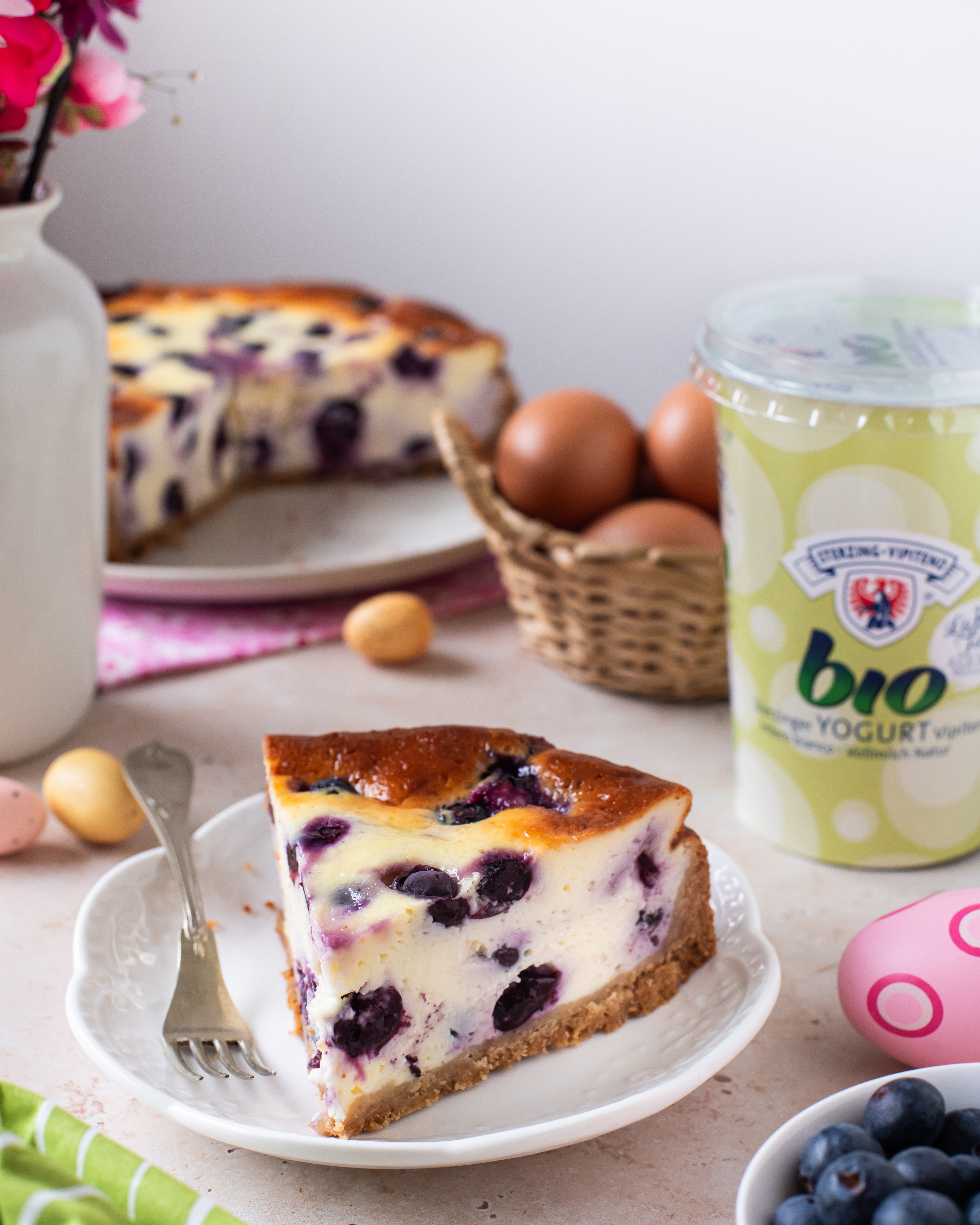 Ein Stück Heidelbeer-Käsekuchen auf weißem Teller mit Gabel, daneben ein Korb mit Eiern, ein Becher Bio-Joghurt und weitere Kuchenstücke im Hintergrund