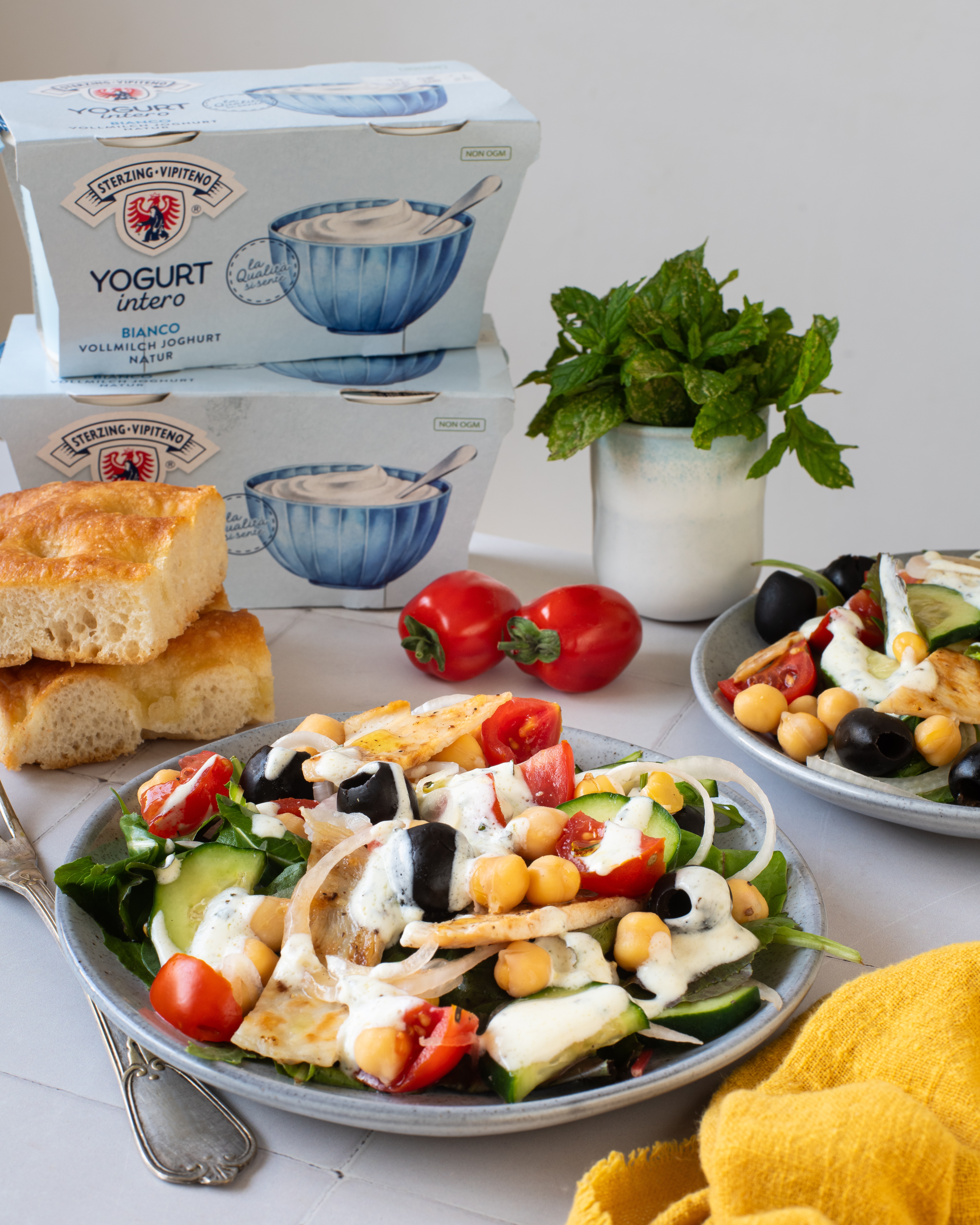 Zwei Teller mit Salat aus Hähnchenstreifen, Kichererbsen, Gurkenscheiben, Tomaten, schwarzen Oliven und Zwiebelringen mit Joghurtdressing, daneben Brot, ein Becher Minze und eine Packung Vollmilchjoghurt