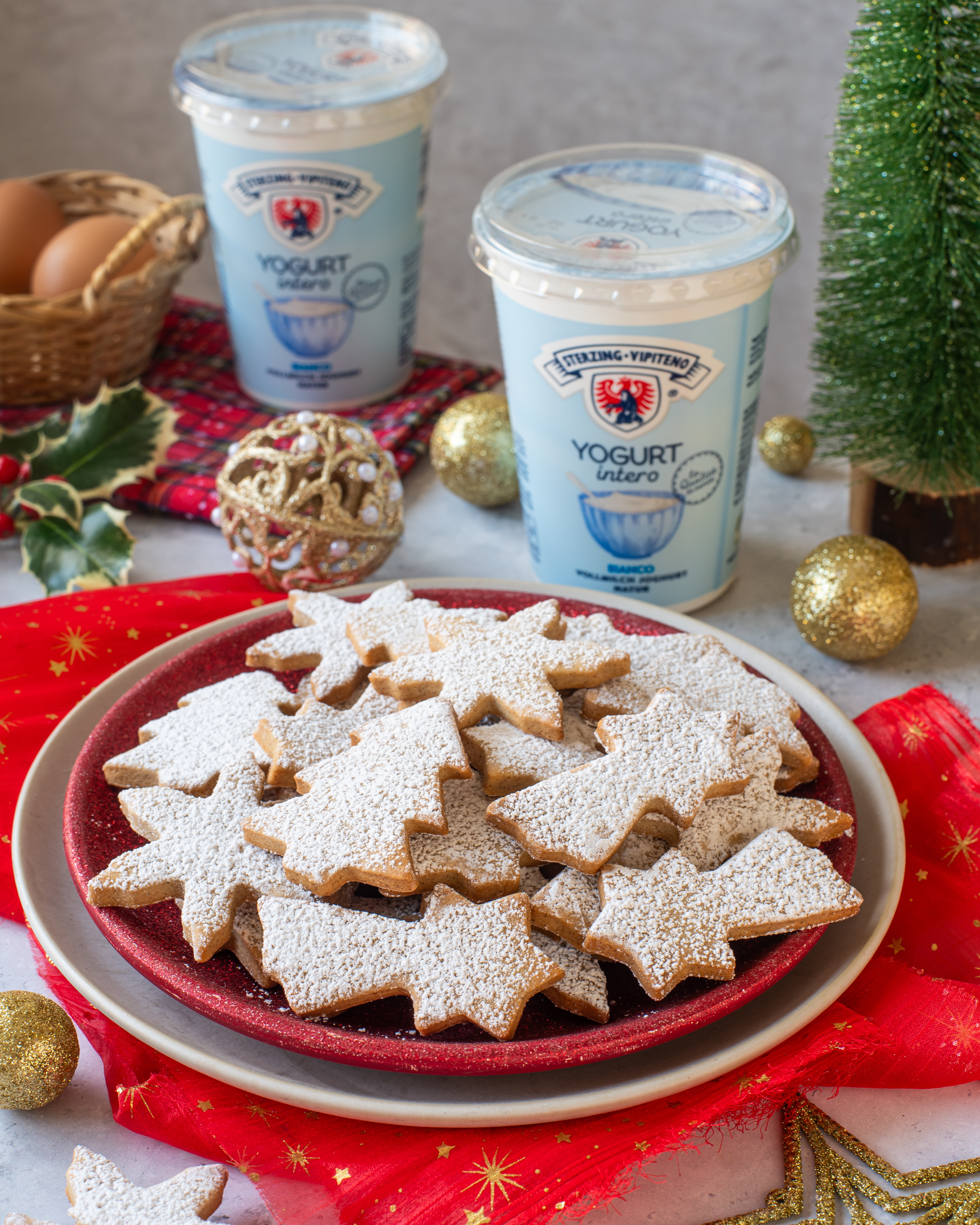 Teller mit stern- und engelförmigen, mit Puderzucker bestäubten Weihnachtsplätzchen neben einem Becher Vollmilchjoghurt und weihnachtlicher Dekoration