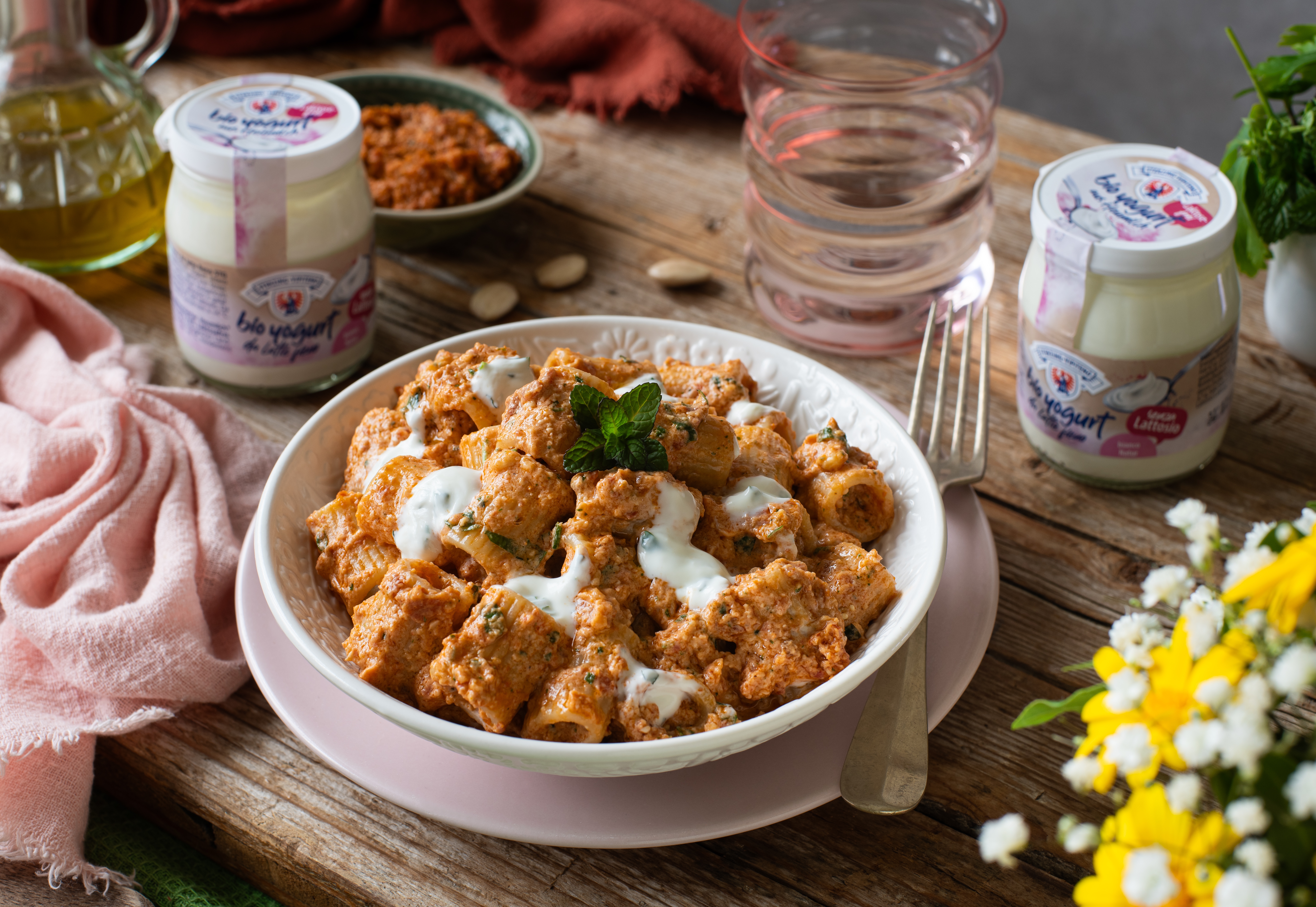 Teller mit Mezze Maniche Pasta in cremiger Pesto-Tomaten-Soße, garniert mit Joghurt und frischen Kräutern auf rustikalem Holztisch mit Joghurtgläsern und Blumen im Hintergrund