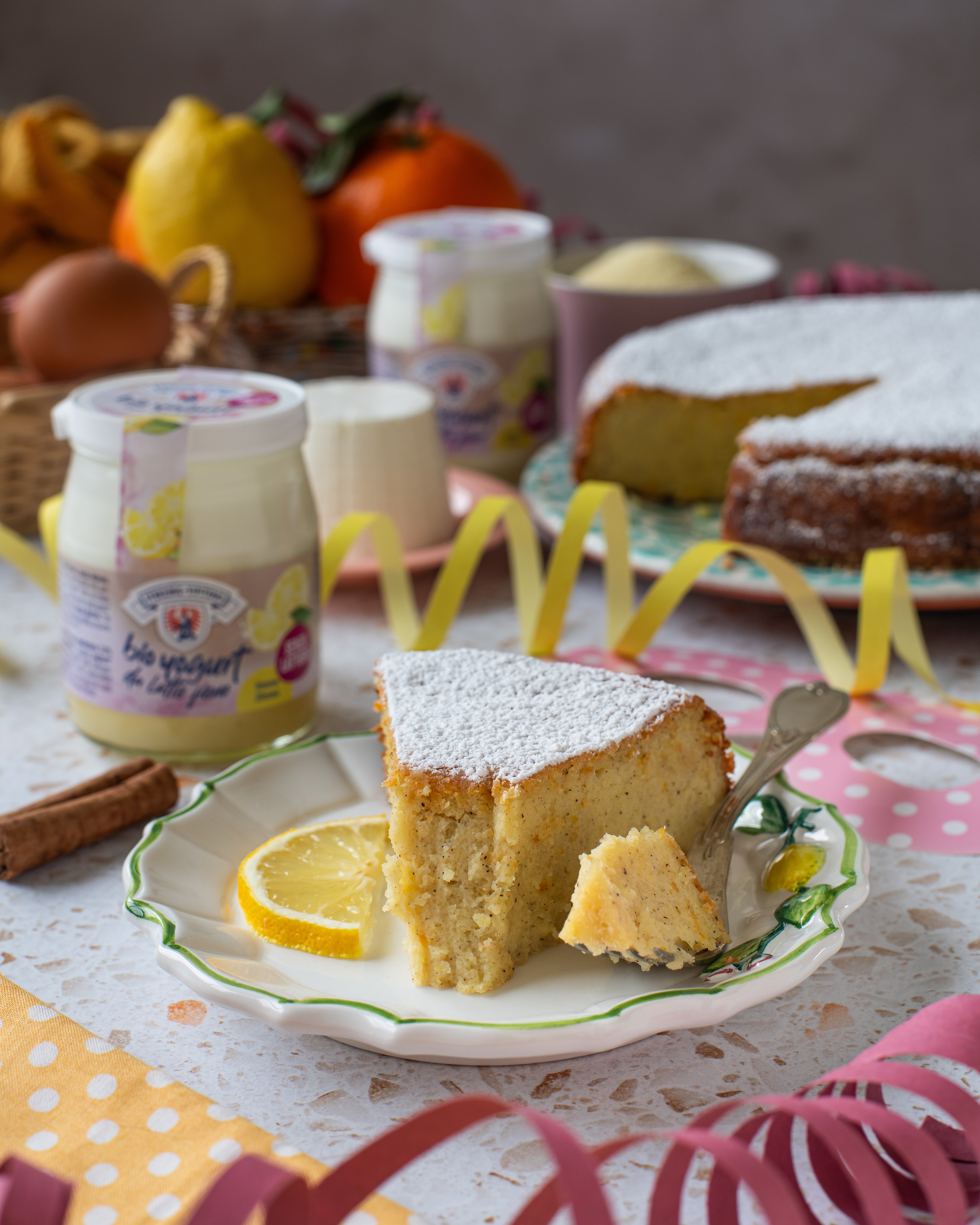 Stück mit Puderzucker bestäubter Kuchen auf Teller mit Zitronenscheiben und Gabel daneben Joghurtgläser, Eierkorb, Zimtstange und Obstkorb auf Tisch mit Tischdecke und Deko