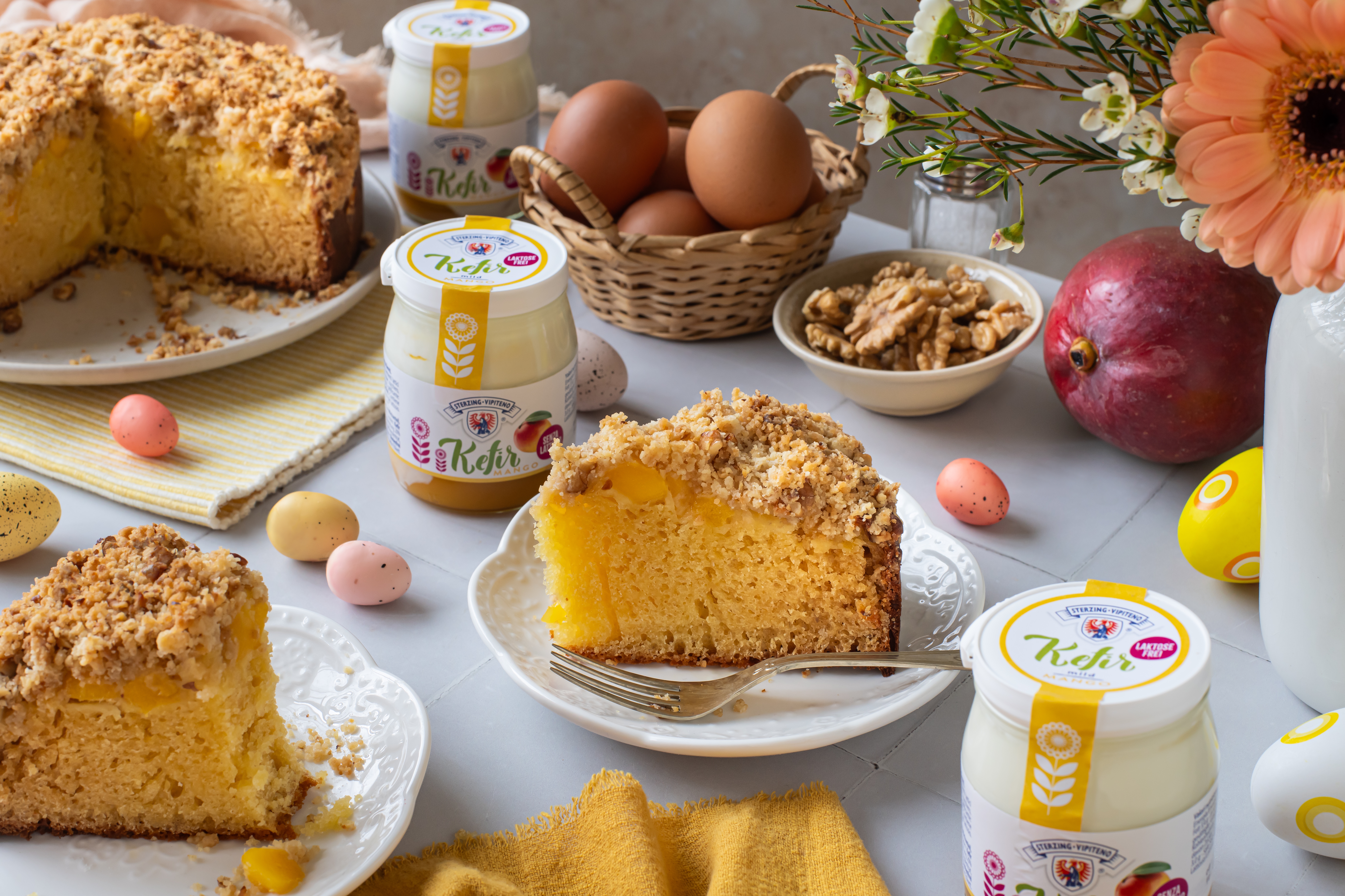 Stück Mango-Crumble-Kuchen auf weißem Teller mit Gabel umgeben von Kefir-Gläsern, Eiern, Walnüssen, Mango und Blumen