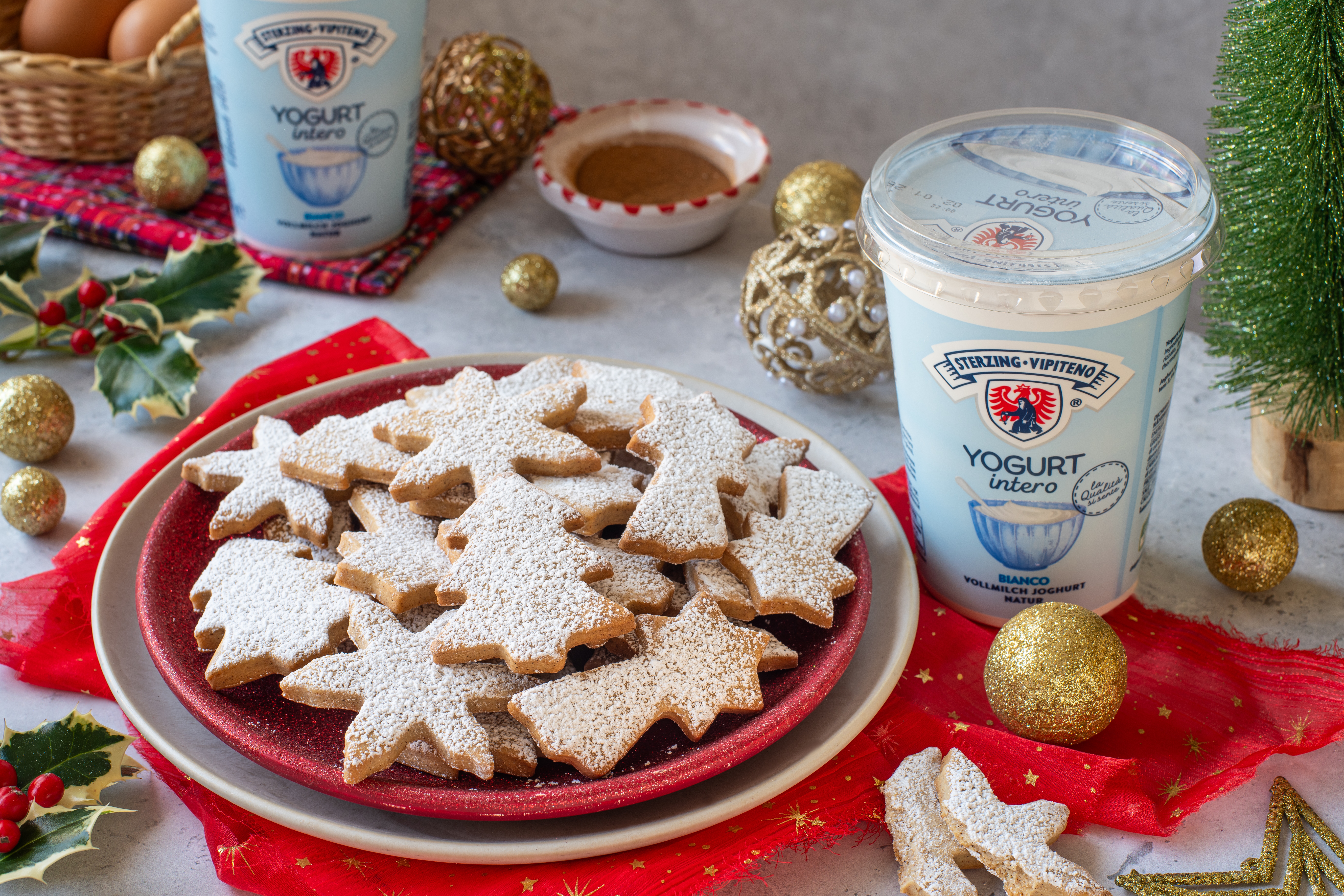 Teller mit stern- und engelförmigen, mit Puderzucker bestäubten Weihnachtsplätzchen neben einem Becher Vollmilchjoghurt und weihnachtlicher Dekoration